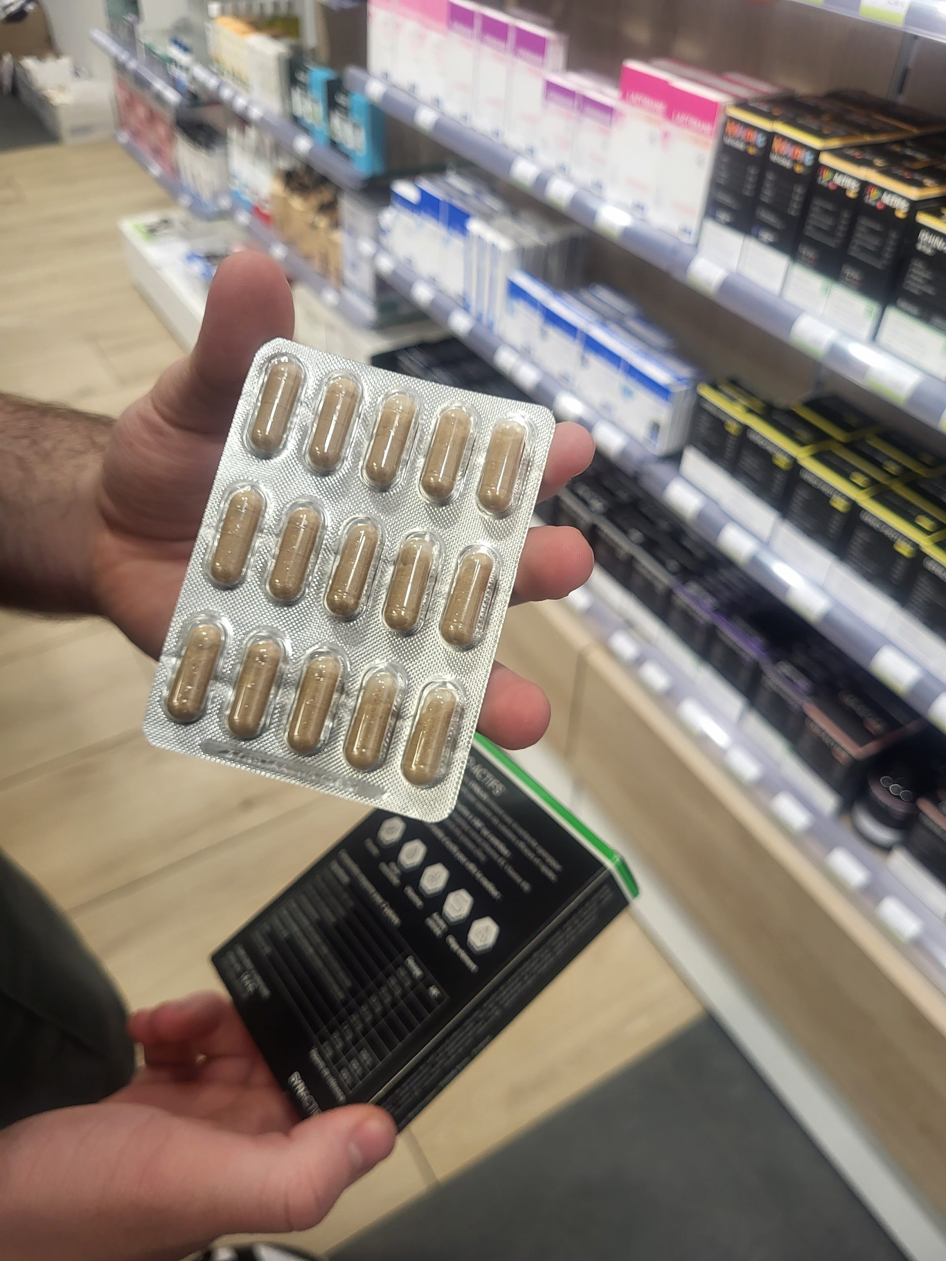 Plaquette de gélules diurétiques tenue en main dans une pharmacie avec des compléments alimentaires en arrière-plan.