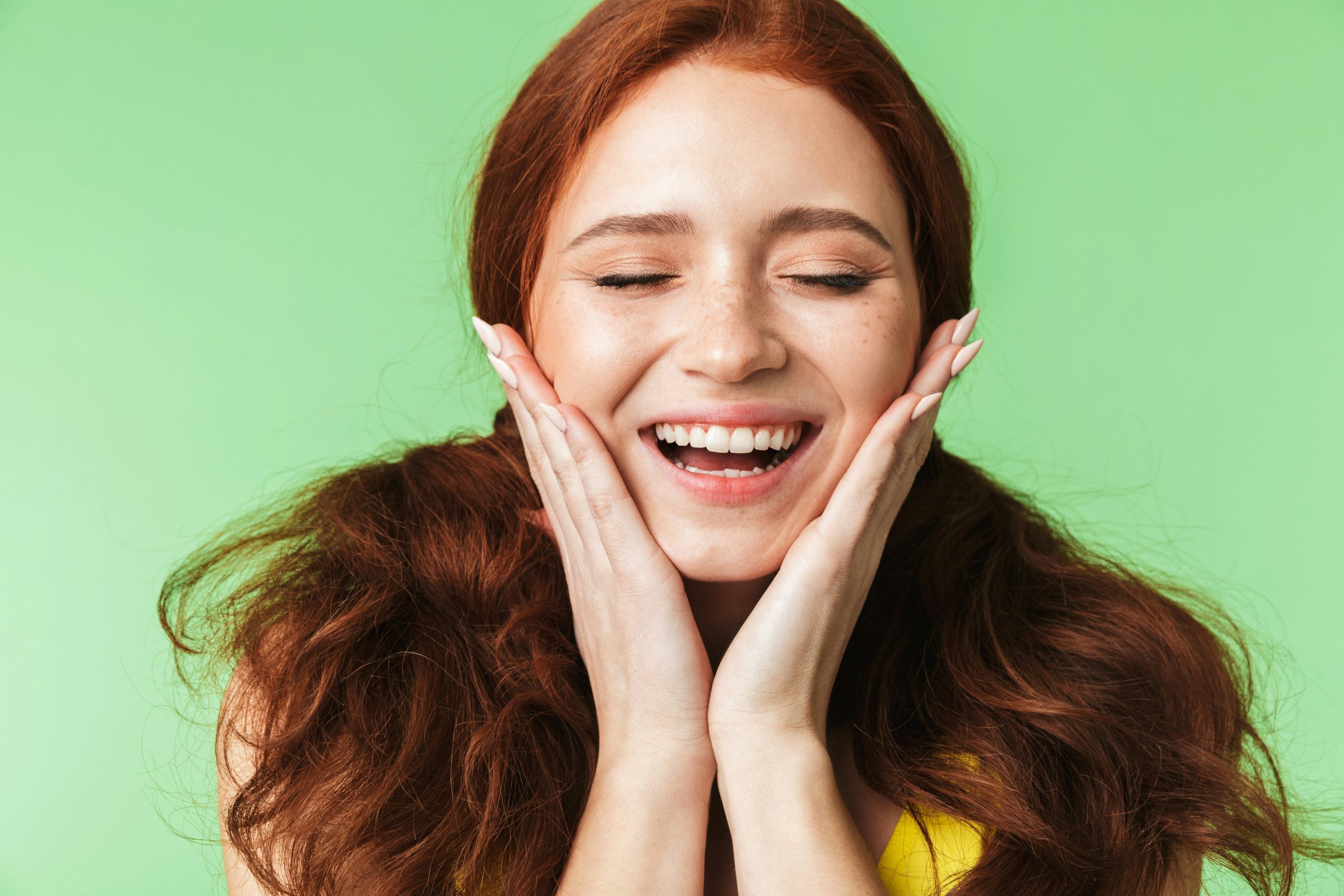 Femme souriante aux dents blanches, posant joyeusement avec les mains sur son visage, sur fond vert.