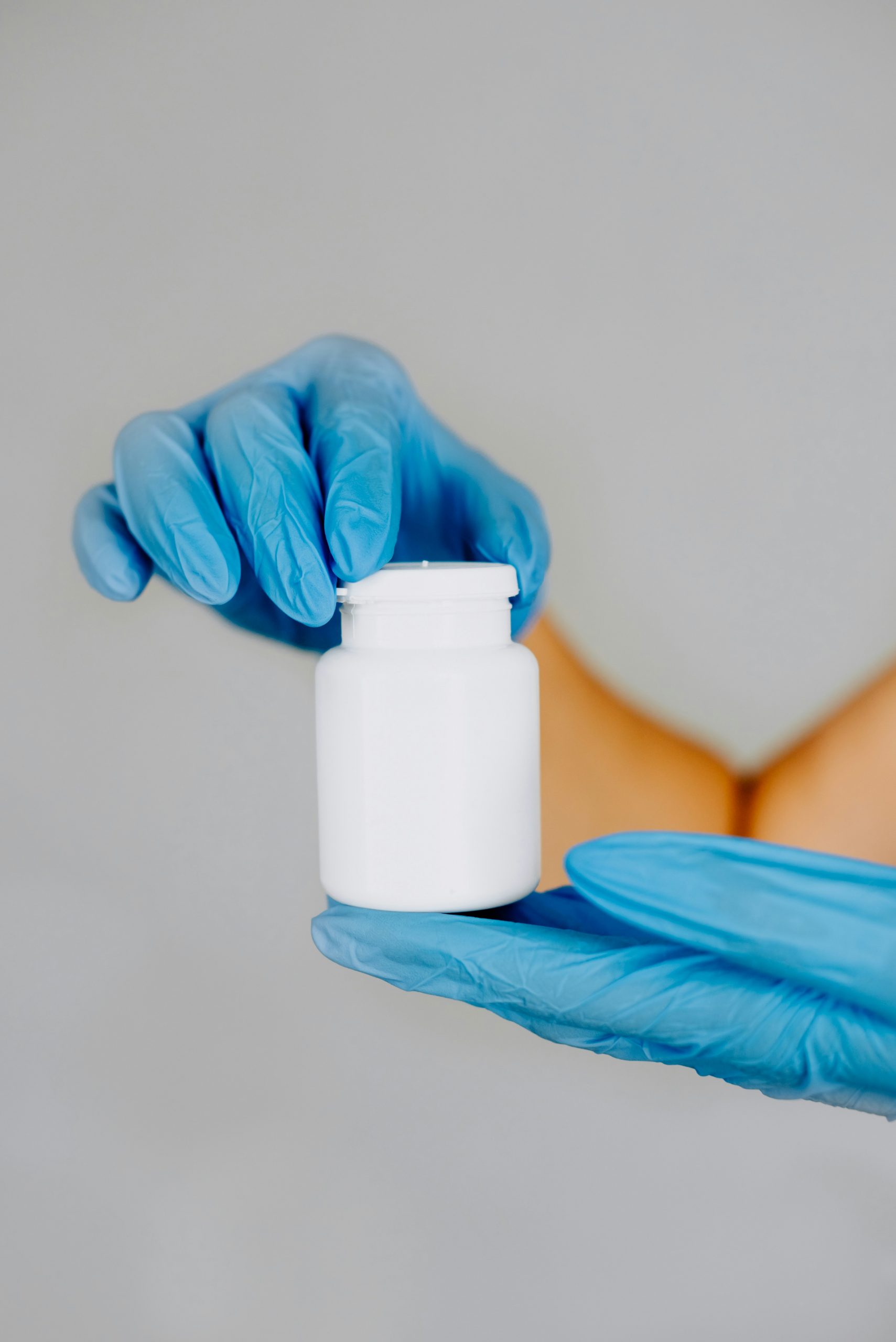 Flacon blanc du médicament Suzetrigine, traitement innovant contre la douleur, tenu par une personne portant des gants médicaux bleus, sur fond neutre.