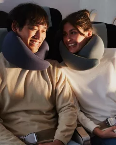 Un couple souriant portant des oreillers de voyage Ostrichpillow Go sur un vol, profitant d’un confort optimal.