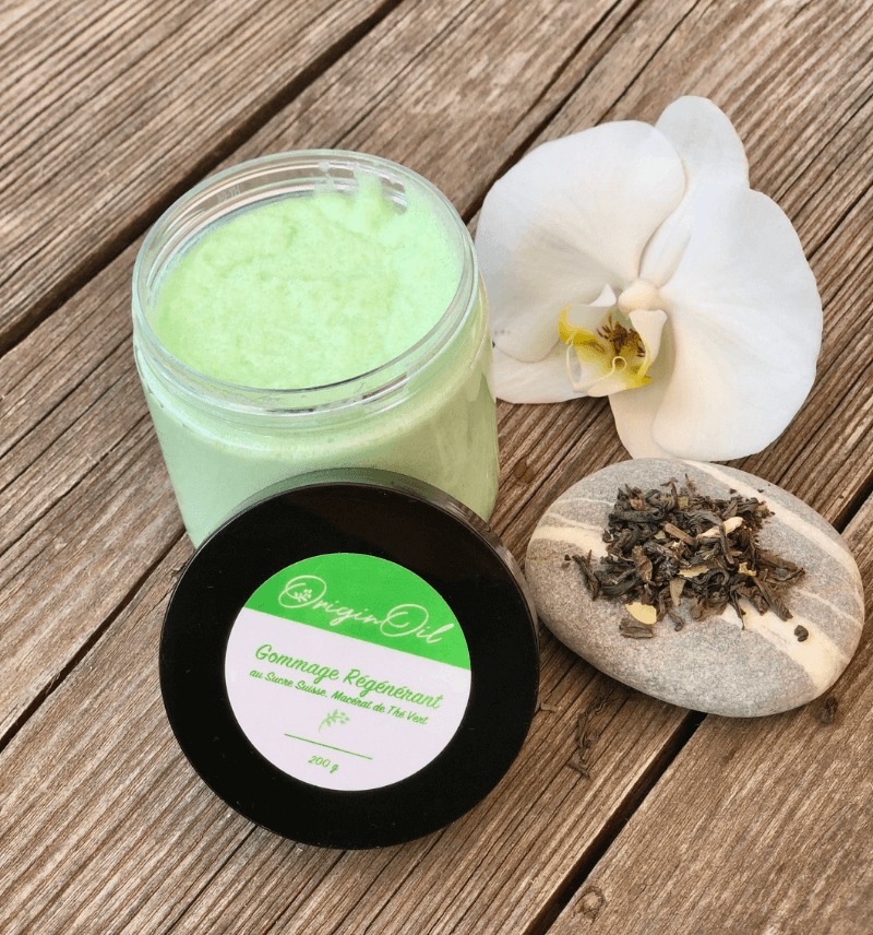 Pot de gommage régénérant vert OriginOil avec sucre cassonade, menthol et thé vert, posé sur une table en bois avec une fleur d’orchidée blanche et des feuilles de thé séchées.